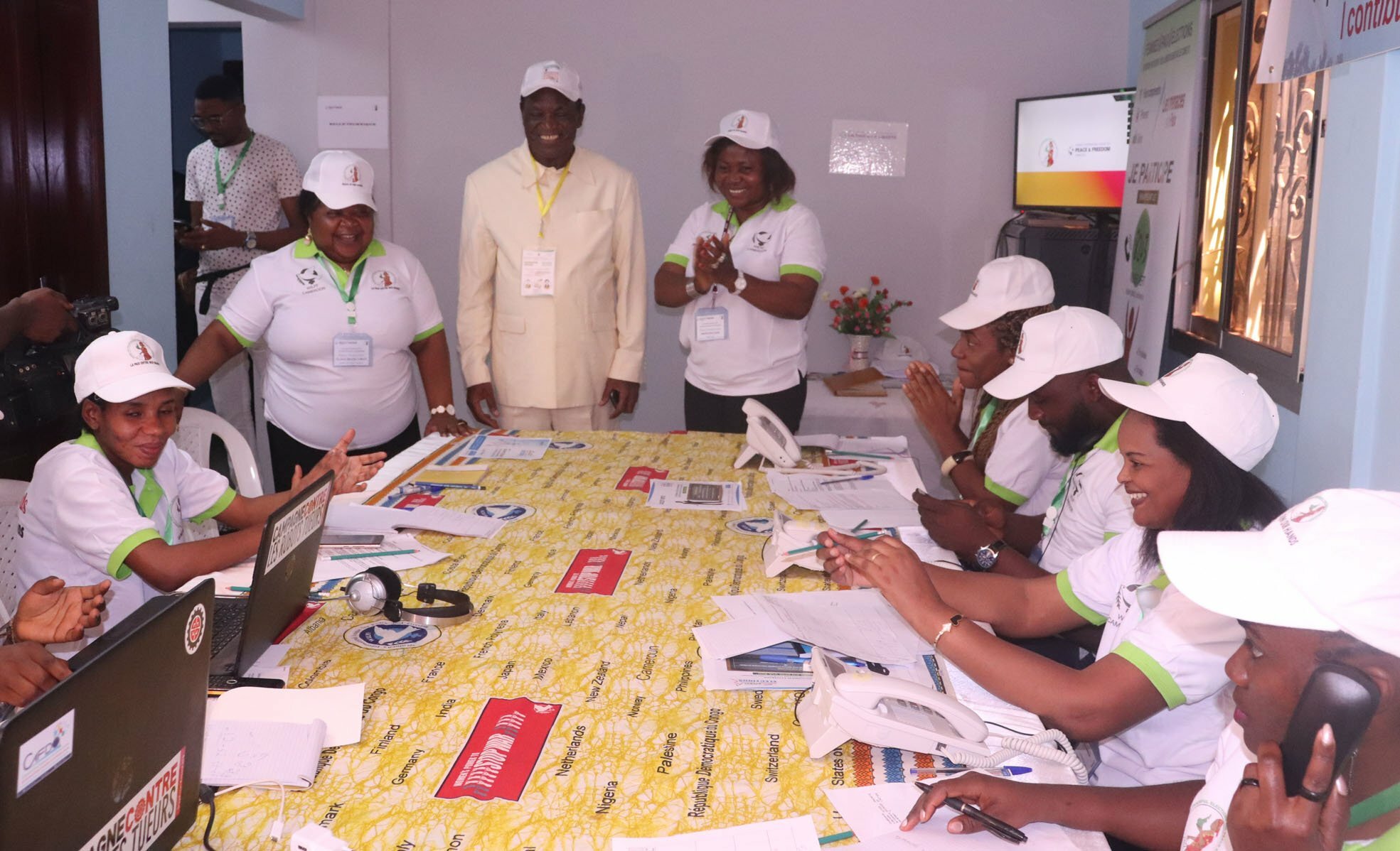 Women working in the Call Center monitoring the violence during the election in Cameroon; they are seated around the table; a woman and a man are standing and smiling while looking at the papers on the table.