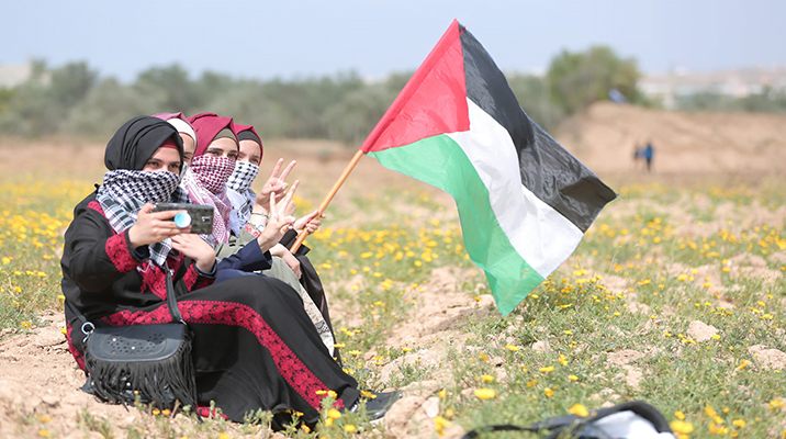 Three girls sitting in a field in Gaza Strip, raising Palestinian flag