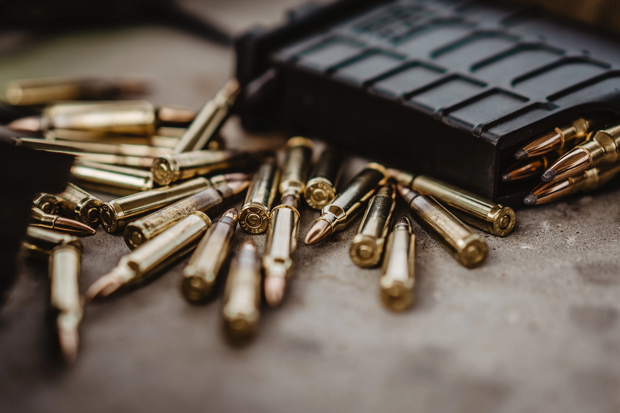 A scattered pile of rifle bullets next to a black magazine on a concrete surface, reflecting light off their metallic casings, serves as a stark reminder of the ongoing journey toward disarmament.