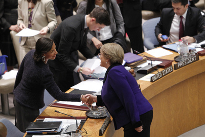 Two women are shaking hands in a formal meeting room setting. One is seated at a desk labeled United States. Papers and officials are visible in the background, indicating an official gathering or conference.