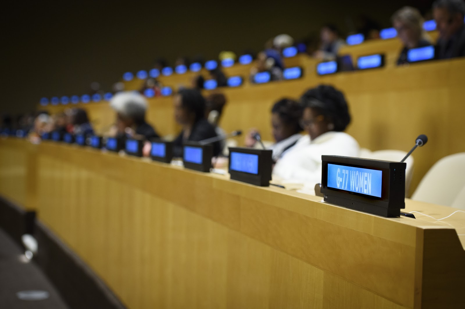 A row of delegates seated at a conference table with digital nameplates in front of them. The focus is on a sign reading G-77 WOMEN. The setting appears to be a formal meeting or conference in a large auditorium.