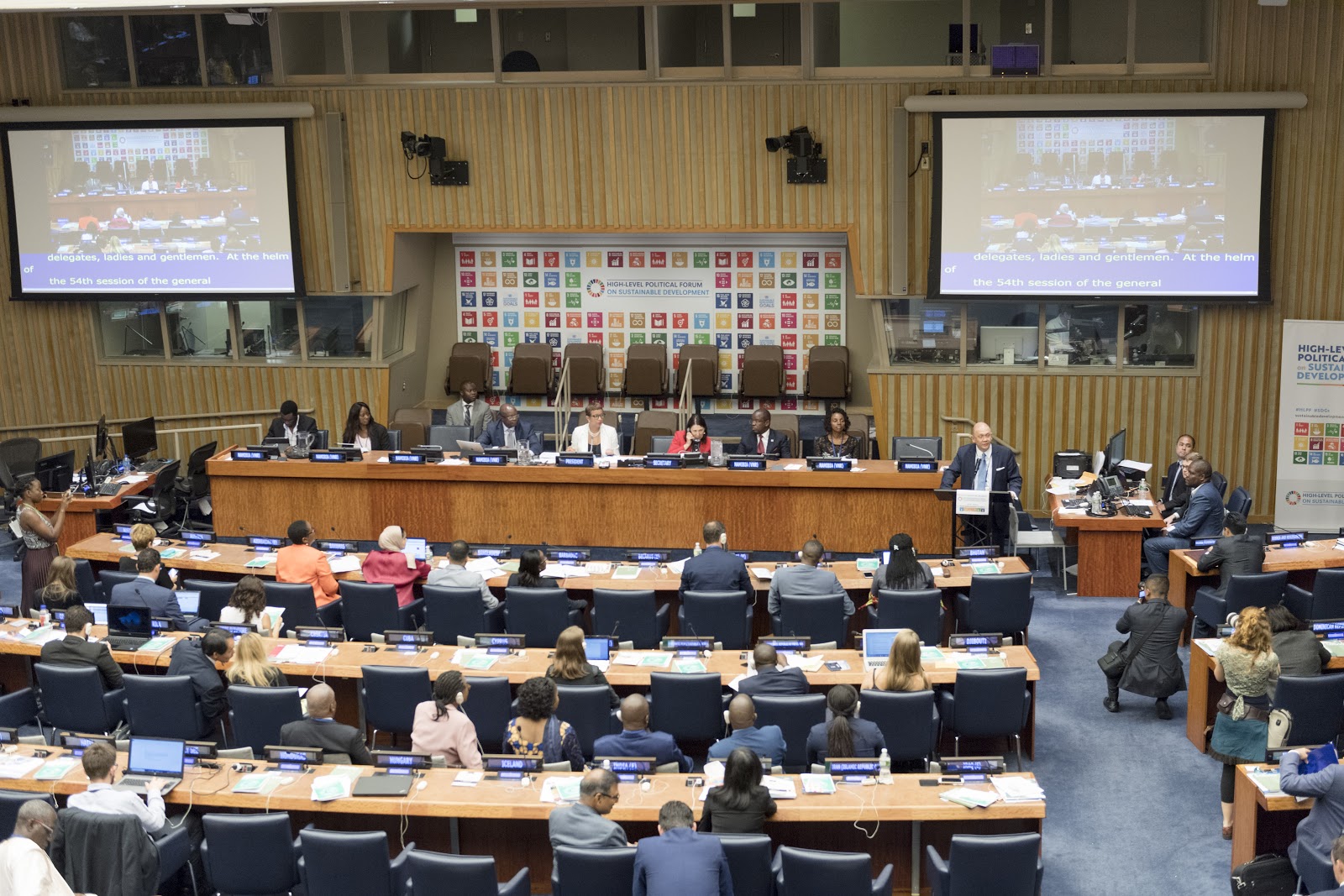A conference room with people seated at desks, facing a long panel of speakers. Two large screens display a presentation. The room is decorated with flags and banners showcasing colorful circular symbols.