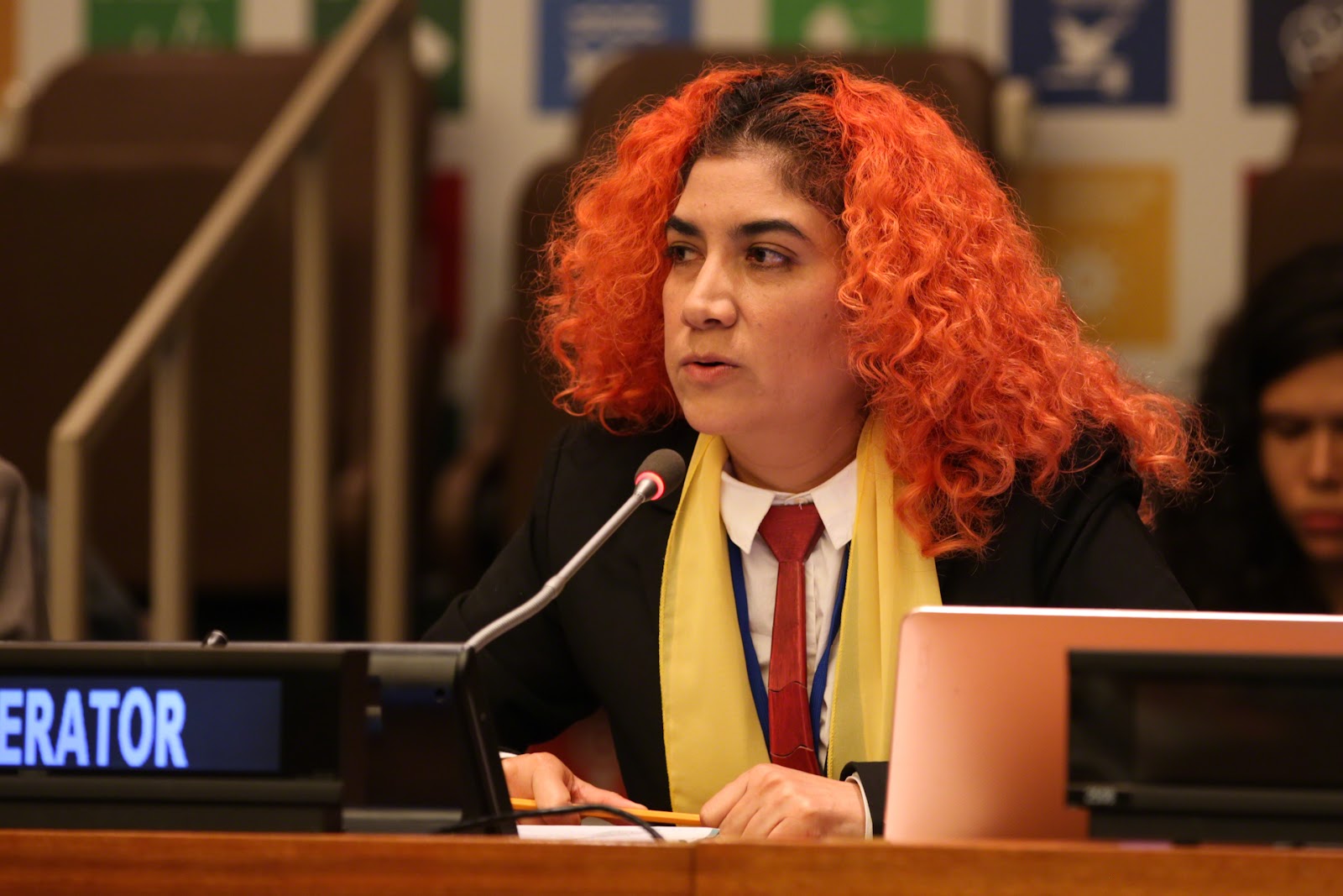 A person with curly, bright red hair speaks into a microphone at a conference. They are wearing a suit with a red tie and yellow scarf. Background shows blurred images and chairs.