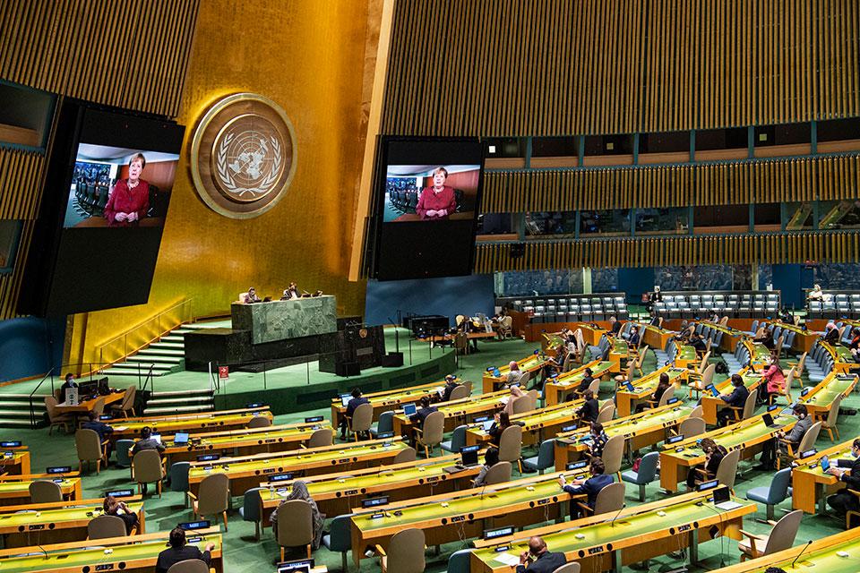 A large, semicircular conference hall with rows of seats facing a central podium. A large emblem and video screens displaying a speaker in red are featured on the back wall. Attendees are seated throughout the hall.