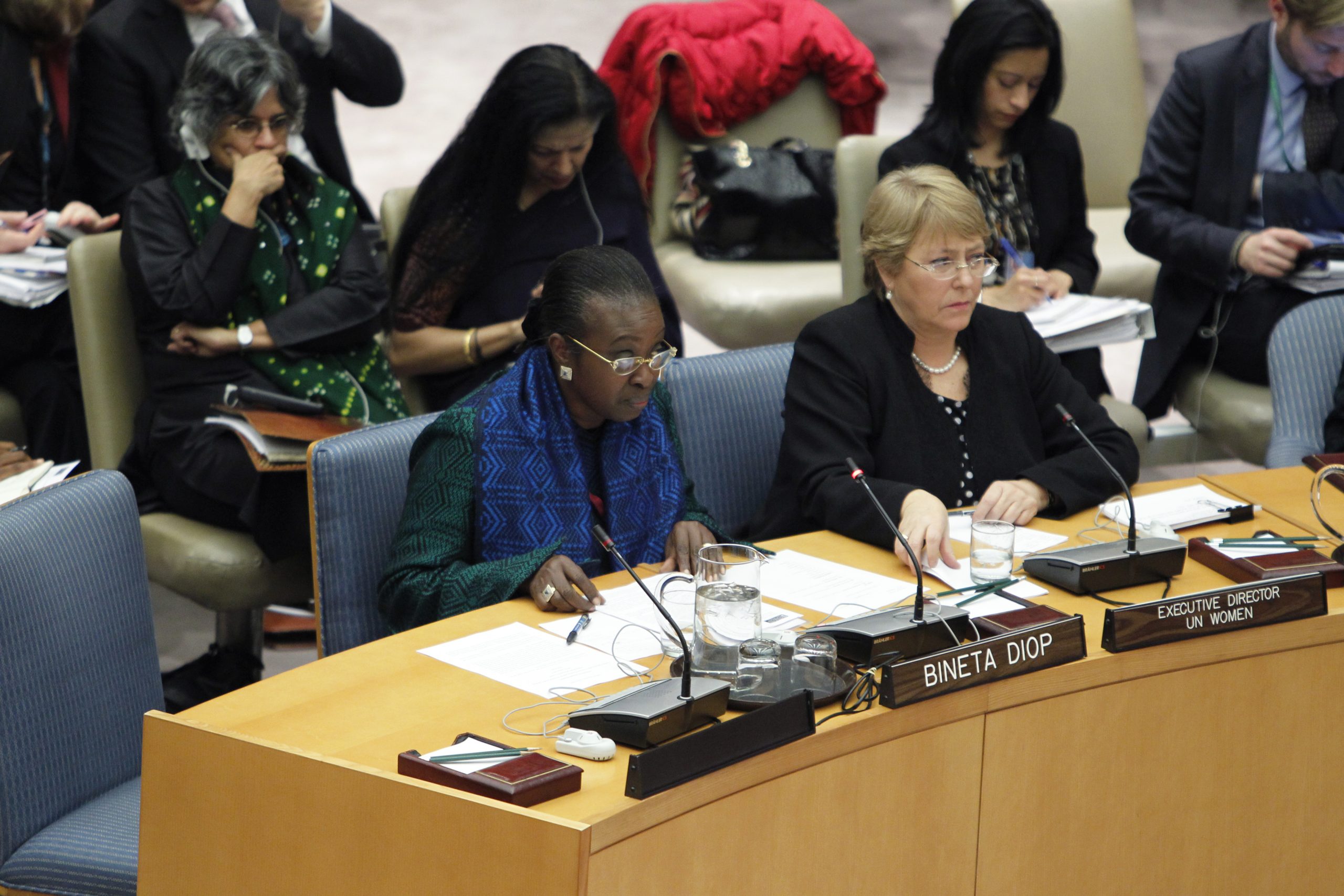 Two women are seated at a conference table with nameplates: Bineta Diop and Executive Director UN Women. They appear engaged in discussion, with papers and microphones in front of them. Other attendees are in the background, listening attentively.