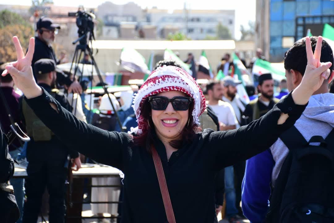 A smiling woman wearing sunglasses and a red-and-white keffiyeh raises both hands in a peace sign at a lively outdoor gathering with people and flags in the background.