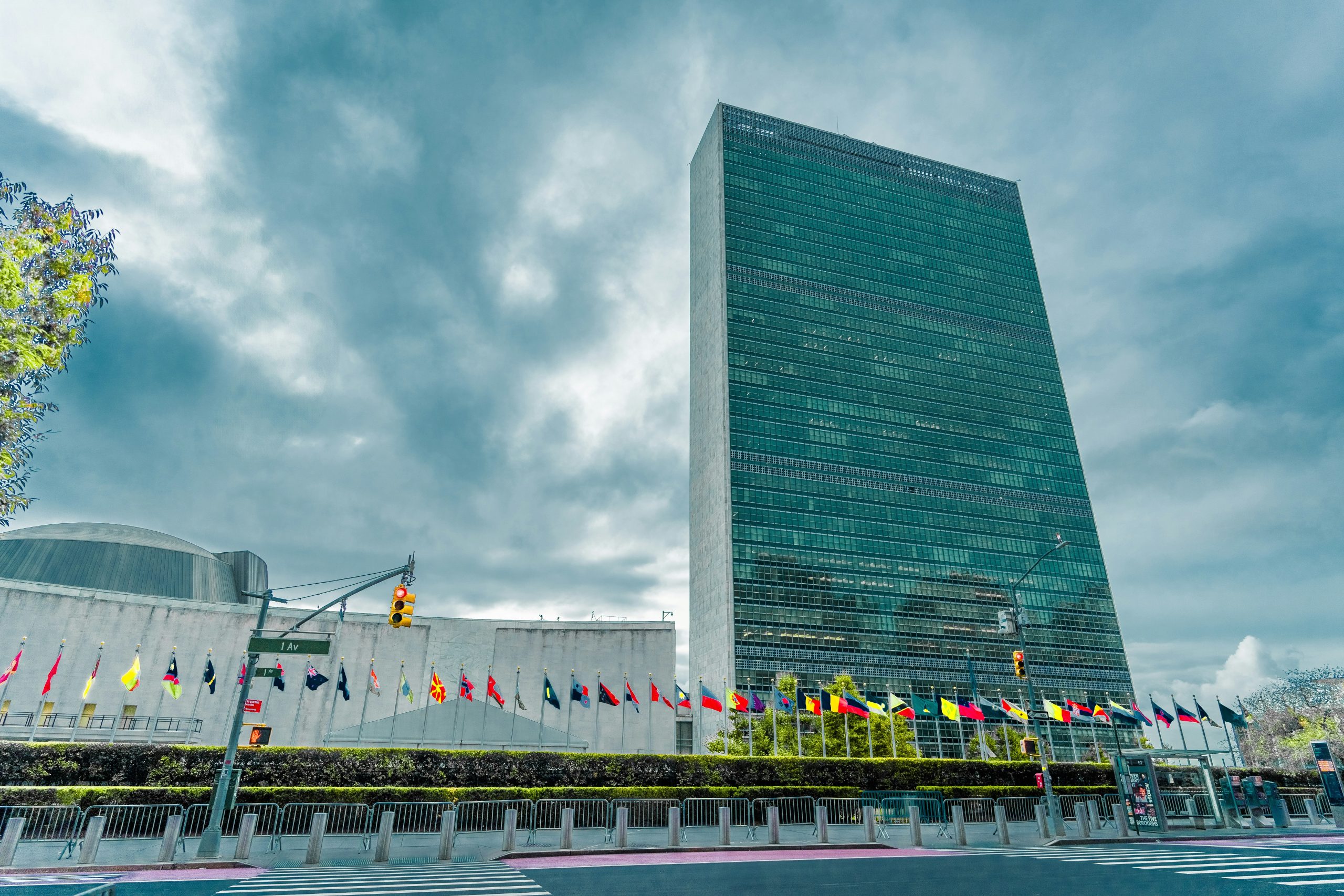 The United Nations Headquarters building in New York City, with rows of international flags displayed outside along the street under a cloudy sky.