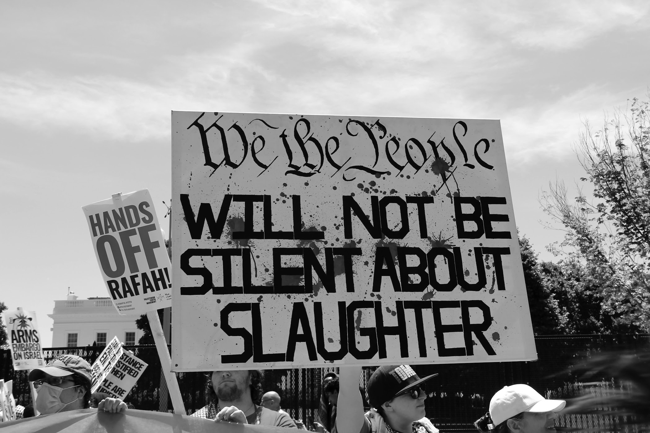 A group of protesters hold signs, including a large one reading “We the People Will Not Be Silent About Slaughter.” Another sign says “Hands Off Rafah!” The protest appears to be outside with trees and buildings visible.