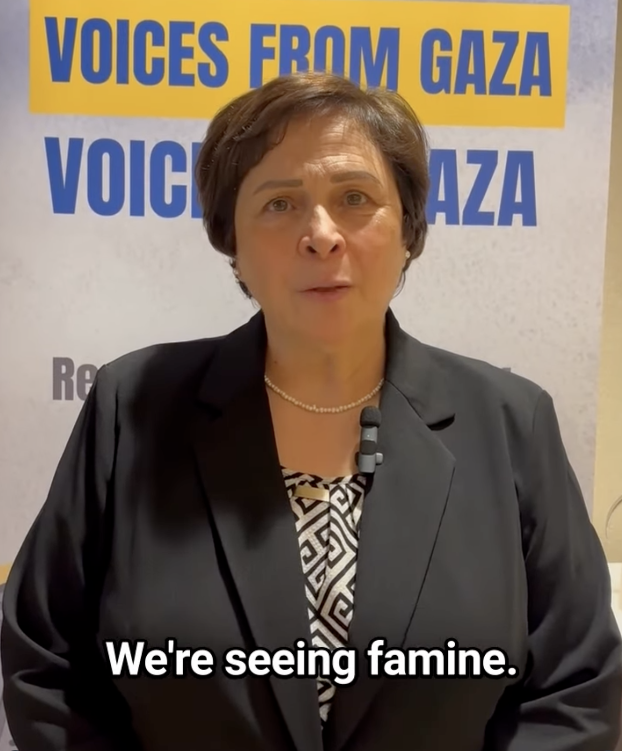 A woman in a black blazer stands in front of a sign that reads Voices from Gaza. She has short hair and speaks toward the camera. Text on the image says, Were seeing famine.