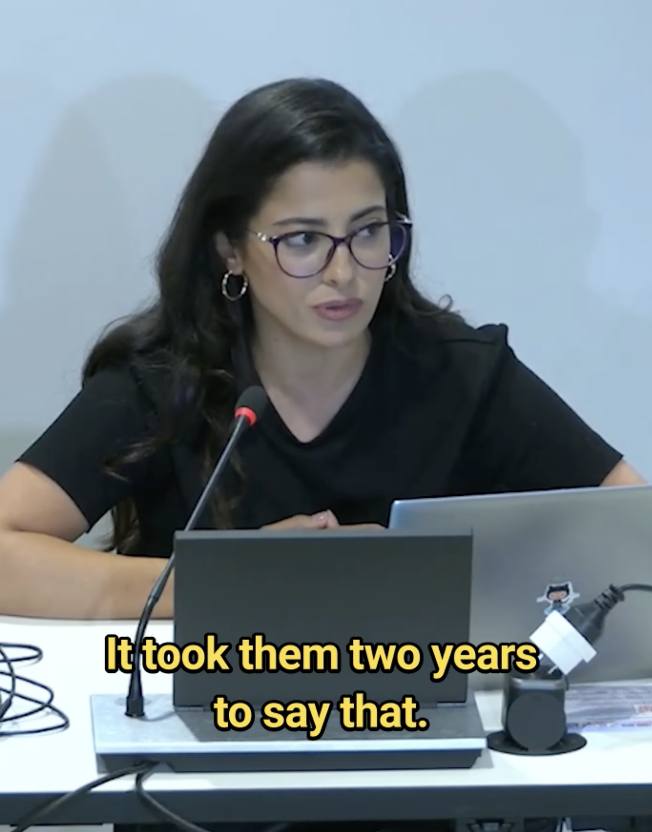 A woman with long dark hair and glasses sits at a table with a laptop and microphone, speaking. Yellow caption text at the bottom reads, It took them two years to say that.