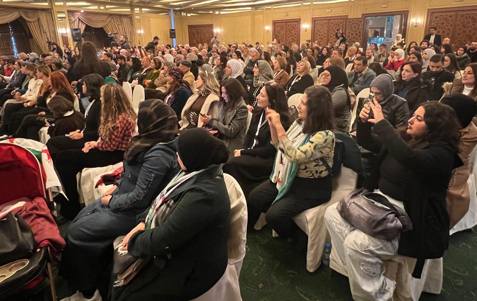 A large audience, mostly women, sit closely together in rows of chairs at an indoor event. Many are wearing headscarves, and several are taking photos or videos with their phones. The room is warmly lit and decorated.