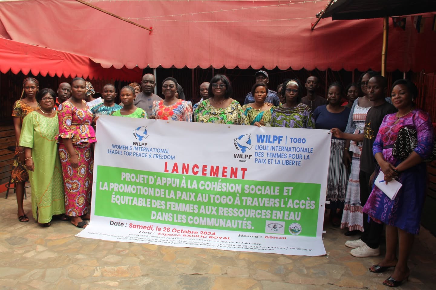 A group of women and men stand together holding a banner in French about launching a project to promote peace and women’s access to water resources in Togolese communities. The event date and details are also visible on the banner.