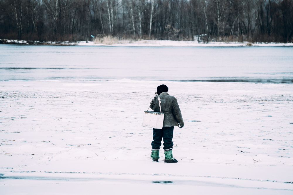 A person dressed in winter clothing stands on a snowy, frozen lakeshore, facing away from the camera and holding a box with a strap, with bare trees visible across the water.