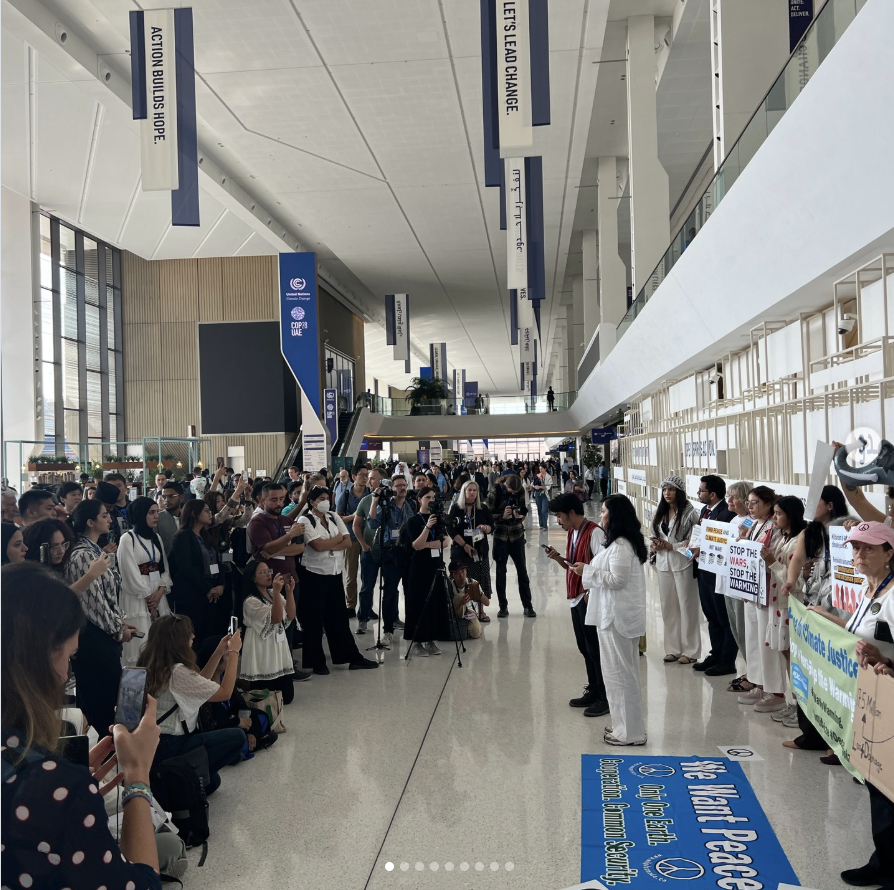 A large group of people gather indoors, some holding placards with messages about peace and justice, while others take photos or watch the scene. The setting appears to be a spacious, modern building with high ceilings.
