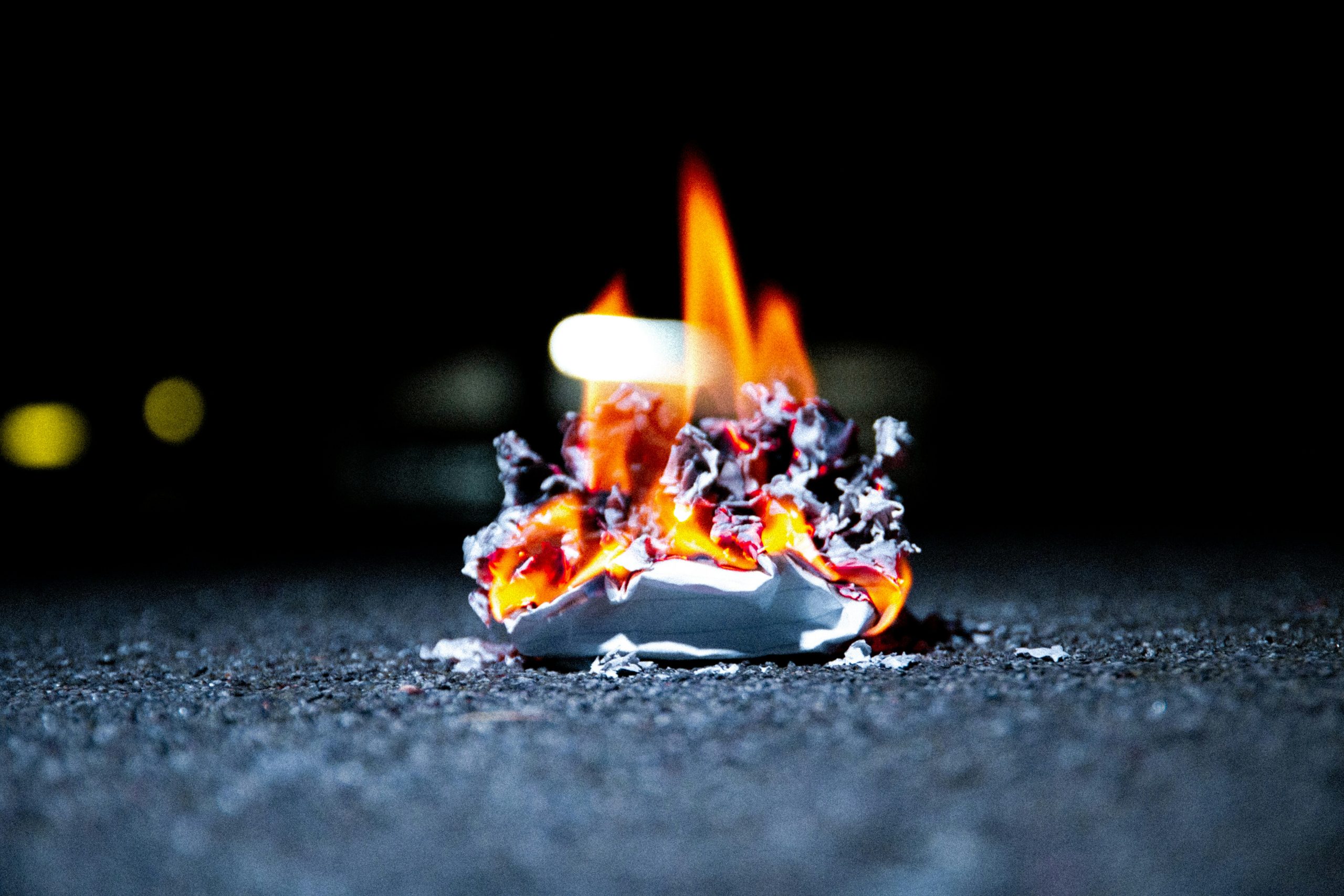 A crumpled piece of paper burns with bright flames on a dark, rough surface, with blurred lights faintly visible in the background.