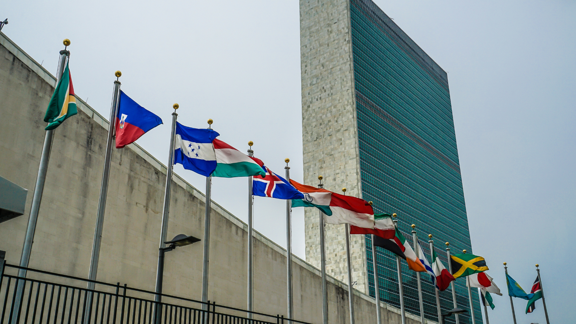 Several national flags are displayed on poles outside the United Nations Headquarters building in New York City, with the distinctive green-glass tower and a cloudy sky in the background.