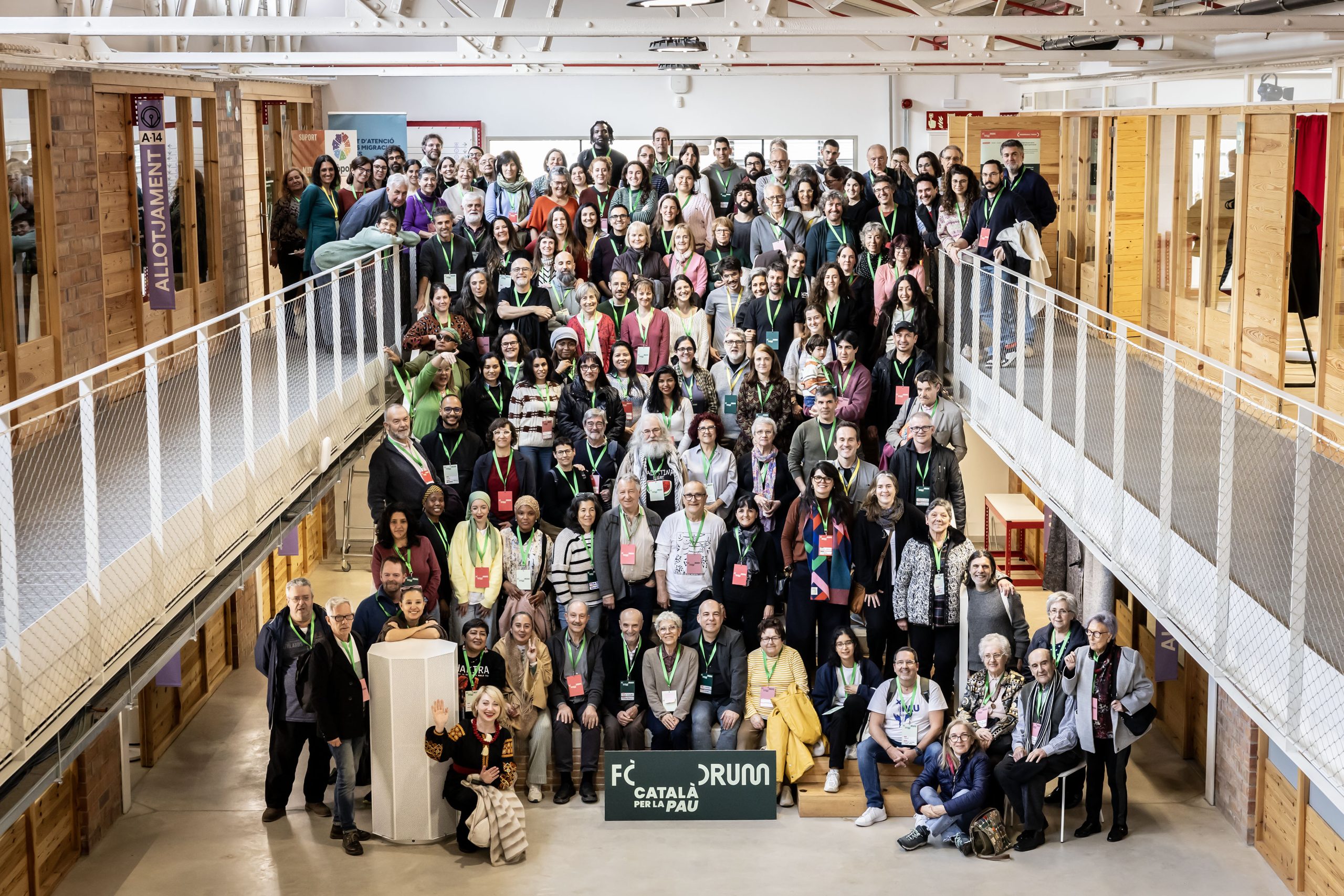 A large, diverse group of people pose together for a group photo inside a modern, wooden building with balconies. Many wear conference lanyards and smile at the camera. A sign in front reads “Fòrum Català per la Pau.”.