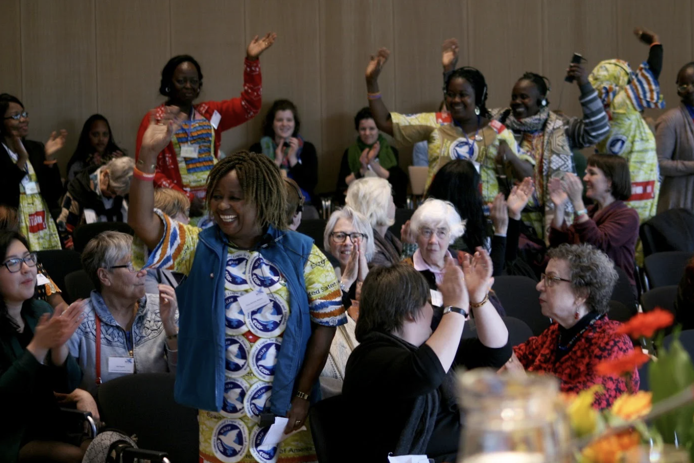 A woman in colourful clothing stands and waves, smiling, as people around her applaud and cheer in a crowded, bright indoor gathering. Others in vibrant attire also wave, celebrating in a lively, joyful atmosphere.