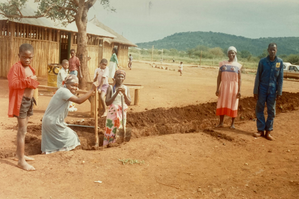 A group of adults and children stand and work by a trench on a dirt ground in a rural area, with wooden buildings and green hills in the background under a cloudy sky.