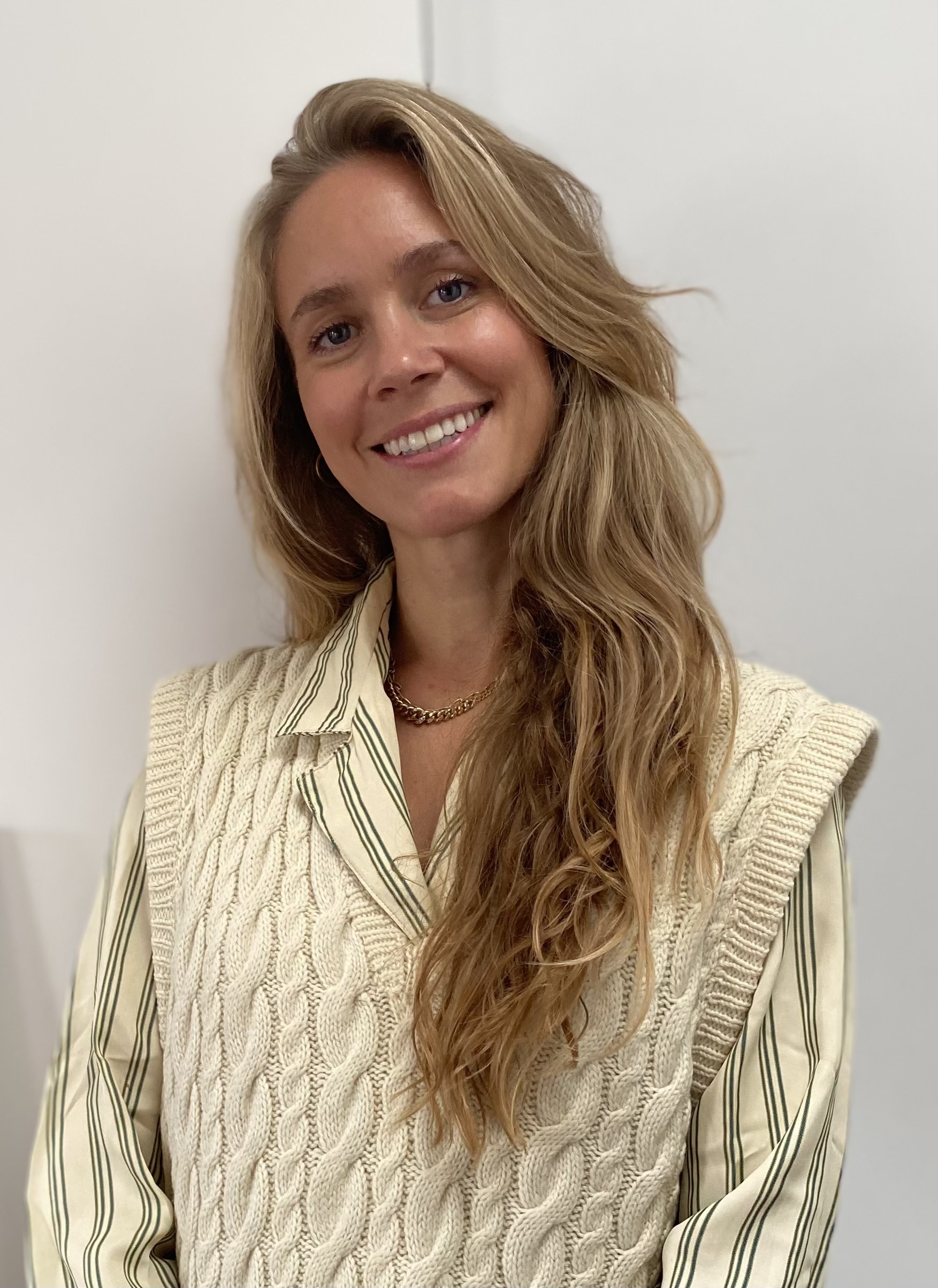 A woman with long, wavy blonde hair smiles at the camera. She is wearing a cream-coloured cable-knit vest over a striped shirt and a gold chain necklace, standing against a plain white background.