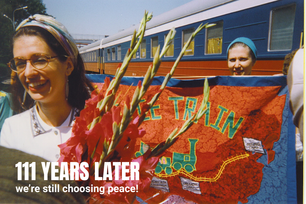 Smiling women hold flowers and a colourful banner with a train and routes, standing beside a train carriage. Overlay text reads, 111 YEARS LATER we’re still choosing peace!.
