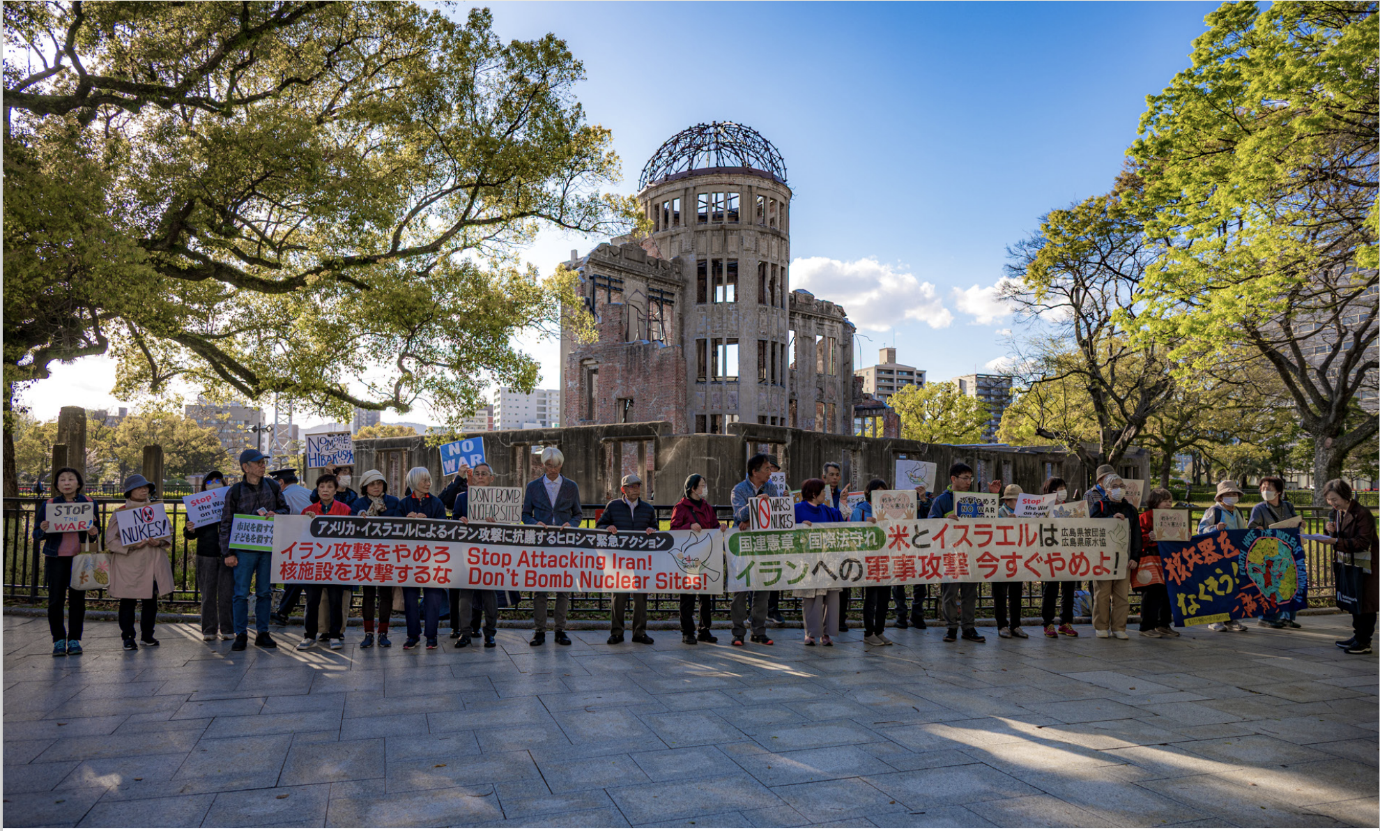 A group of people stand in front of the Hiroshima Peace Memorial holding colourful banners and signs protesting nuclear war and military action, with trees and the ruined dome in the background.