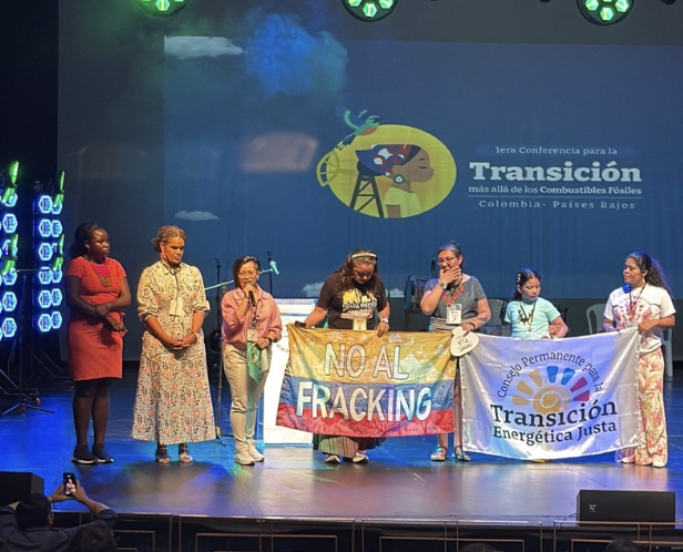A group of women stand on stage at a conference, two holding banners that read “NO AL FRACKING” and “Transición Energética Justa.” Stage lights shine behind them, and a conference sign is visible in the background.
