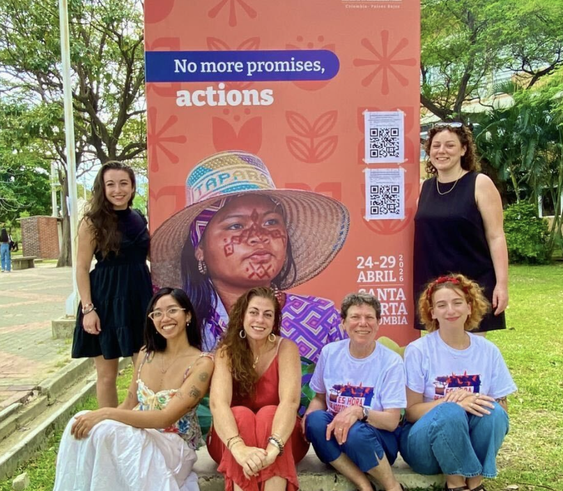 Six women pose and smile in front of a large colourful poster that reads, No more promises, actions, with event details and QR codes. Trees and greenery surround the group in an outdoor setting.